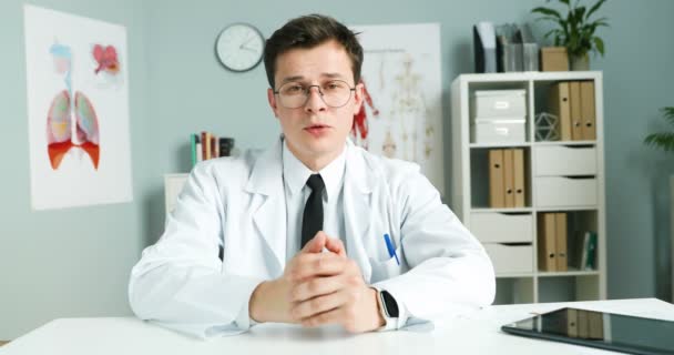 Hombre doctor blogger en bata blanca y gafas sentado en el escritorio en el gabinete y contando sobre la atención médica en línea. Blogging on health. Médico que tiene consulta en Internet. Consultoría médica en vlog
. - Imágenes, Vídeo