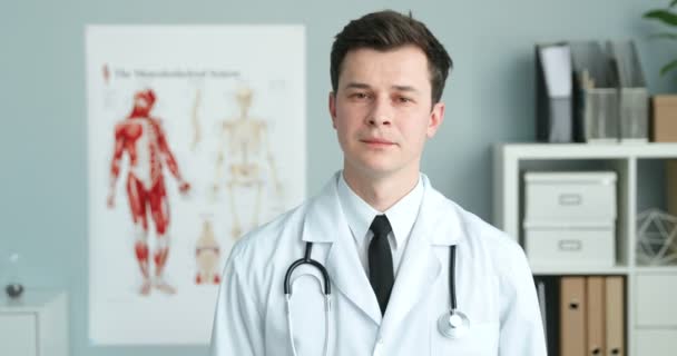 Portretfoto van knappe jonge blanke man in witte jurk en met stethoscoop in zijn kast en glimlachend naar de camera. Goed uitziende mannelijke dokter. - Video