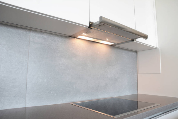 Κοντινό πλάνο στο κάτω μέρος του ντουλαπιού, απορροφητήρα εξάτμισης κουκούλα με φώτα εργασίας και σύγχρονη ηλεκτρική κουζίνα, εστία μαγειρέματος σε γκρι και λευκό σχεδιασμό κουζίνας. - Φωτογραφία, εικόνα