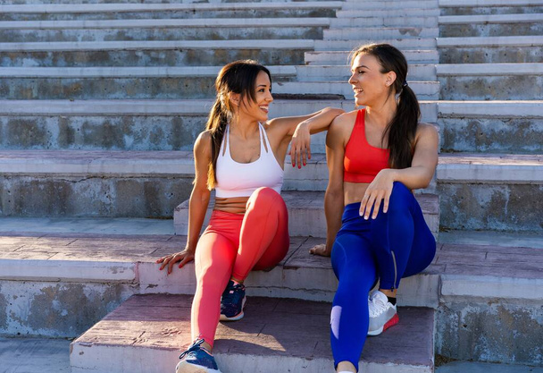Kaksi nuorta valkoihoista urheilullista naispuolista ystävää istuu portaissa ja puhuu kovan harjoittelun jälkeen. - Valokuva, kuva