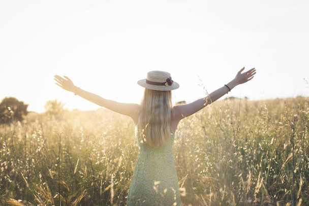 A beautiful shot of a blonde young female wearing a hat standing in the field at sunset - Fotografie, Obrázek