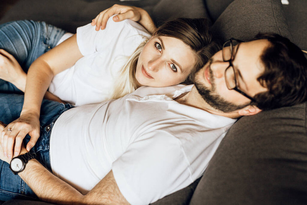 白いTシャツを着た若いカップルのトップビュー。ソファの上で抱擁で幸せな家族。女はカメラを見て - 写真・画像