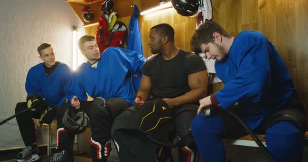 Joueurs de hockey masculins de races mixtes assis dans la cabine d'essayage et parlant après le match. Des sportifs multiethniques discutent et vérifient leur équipement dans le vestiaire pendant la pause. - Séquence, vidéo