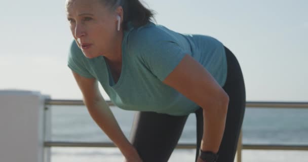 Senior Caucasian woman working out on promenade by the sea wearing sports clothes, taking break with earphones on in slow motion. Retirement healthy lifestyle activity. - Materiaali, video