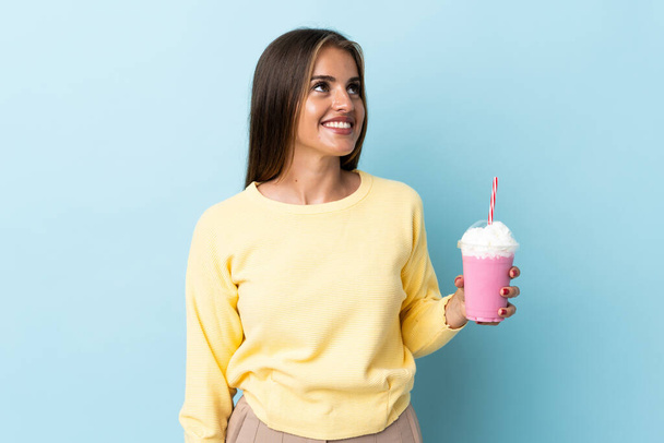 Jonge Uruguayaanse vrouw met aardbeien milkshake geïsoleerd op blauwe achtergrond denken een idee terwijl op zoek omhoog - Foto, afbeelding