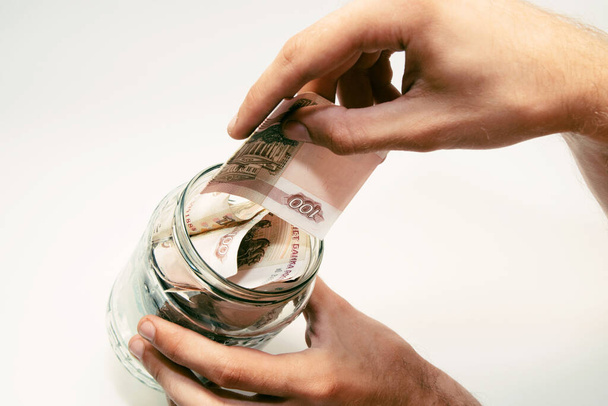 A man holds a glass jar filled with banknotes and puts money in it. - Фото, изображение