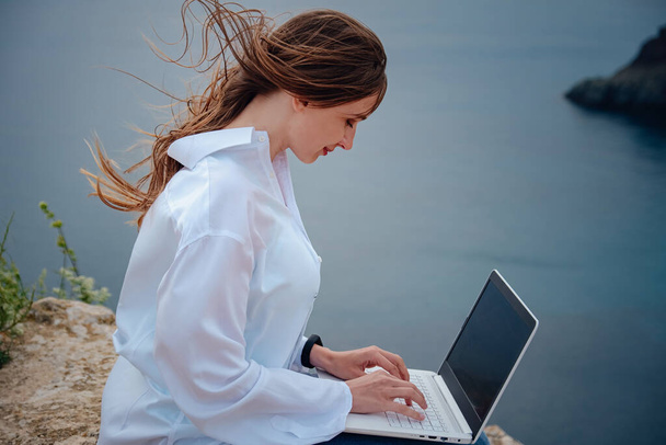 岩の表面の浜のコンピュータで働く若いビジネス女性。海に山の上のノートパソコンで働いている若い女の子、仕事の日. - 写真・画像