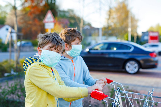 Два мальчика в медицинской маске в качестве защиты от пандемии коронавируса. Дети, использующие защитное оборудование против ковида 19 и идущие за покупками в супермаркет с тележкой. - Фото, изображение