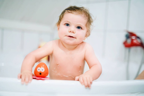Cute adorable baby girl taking foamy bath in bathtub. Toddler playing with bath rubber toys. Beautiful child having fun with colorful gum toys and foam bubbles - Zdjęcie, obraz