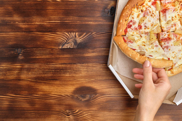 Hawaiianische Pizza in Kartonverpackung und Hand nimmt ein Stück davon, um Platz zu kopieren. - Foto, Bild