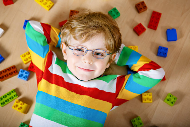 Klein blond kind met bril spelen met veel kleurrijke plastic blokken binnenshuis. Kid jongen het dragen van kleurrijke shirt en plezier hebben met het bouwen en maken - Foto, afbeelding