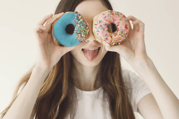 Funny portrait of a young European female model holding blue and pink donuts near eyes and showing a tongue. Positive and cheerful mood - Фото, изображение