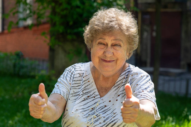 Щаслива стара жінка показує великий палець. Сидить на вулиці в сонячний літній день
. - Фото, зображення