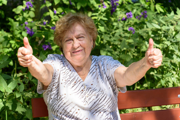 Gelukkige oudere vrouw die duimen laat zien. Zit op een bankje in het park. - Foto, afbeelding