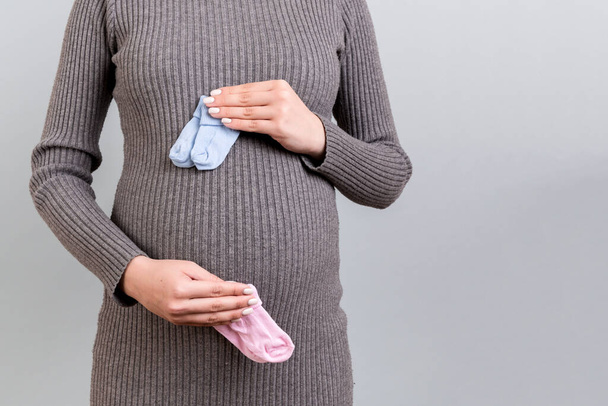 Close up of pregnant woman in gray dress holding baby pink and blue socks against her belly at gray background. Expecting twins. Is it a boy or a girl Child expecting concept. Copy space. - Fotografie, Obrázek