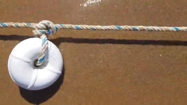 White sea buoy on rough nautical rope knot is waving by clear water sea surf. Marine buoy and rope on wet sea sand background with copy space. Beach summer vacation. Coastal themed backdrop - Footage, Video