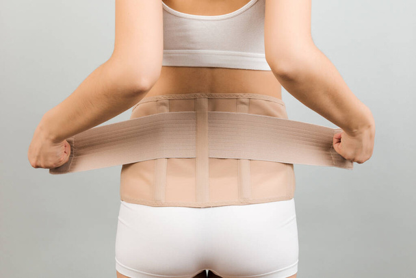Back view of pregnant woman in underwear dressing orthopedic corset to make the backache go away at gray background with copy space. Close up of orthopedic abdominal support belt concept. - Photo, Image