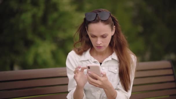 Pretty brunette woman is texting message on cell phone while sitting on a bench in the park. Outdoors portrait - Imágenes, Vídeo