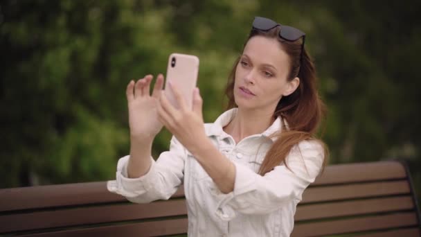 Pretty woman is taking selfie on her smartphone in the park. Summer day. Outdoors portrait - Metraje, vídeo