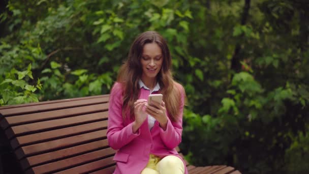 beautiful young woman is wearing bright clothes sitting on a bench in the park and talking on a smartphone - Filmati, video