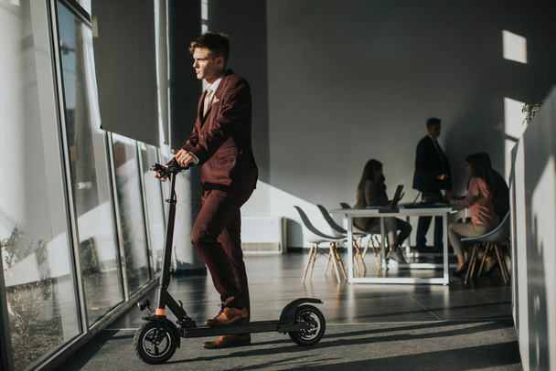 Knappe jonge zakenman bij het raam van het kantoor met elektrische scooter - Foto, afbeelding