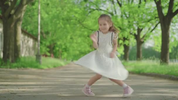 Cheerful Caucasian girl spinning and jumping in sunny summer park. Wide shot portrait of pretty little brunette child enjoying good weather outdoors. Leisure, lifestyle, childhood, happiness. - Metraje, vídeo
