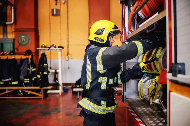 Bombero en uniforme protector con casco en el control de la cabeza en las mangueras antes de la intervención mientras está de pie en la estación de bomberos
. - Foto, imagen