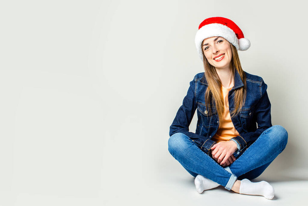 Улыбающаяся молодая девушка, сидящая в шляпе Санта Клауса на светлом фоне
 - Фото, изображение