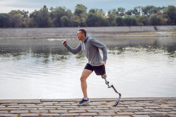 Oldalnézet egészséges kaukázusi fogyatékos sportoló sportruházatban és mesterséges láb fut a rakparton. - Fotó, kép