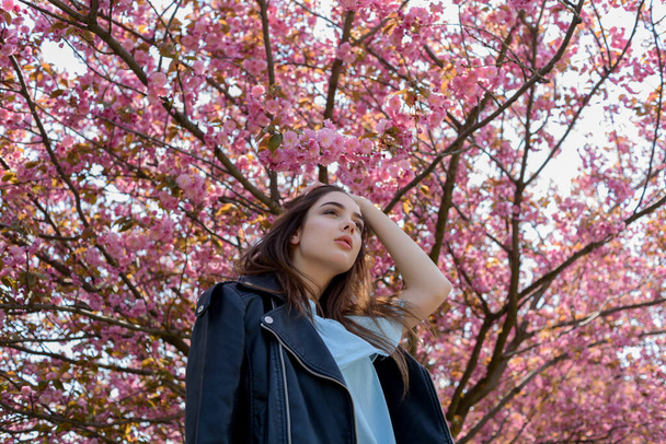 長い髪の若い美少女は、開花桜の木の近くの春の自然の美しさを楽しんでいます。さくらの革のジャケットの女の子. - 写真・画像