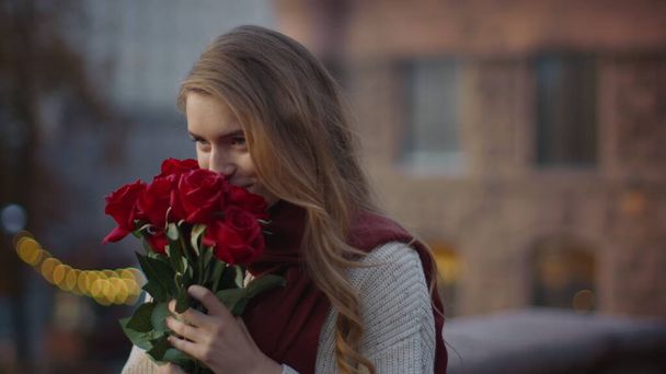 Suloinen tyttö haistaa kukkakimpun ulkona. Söpö nainen kantaa ruusuja kadulla - Valokuva, kuva