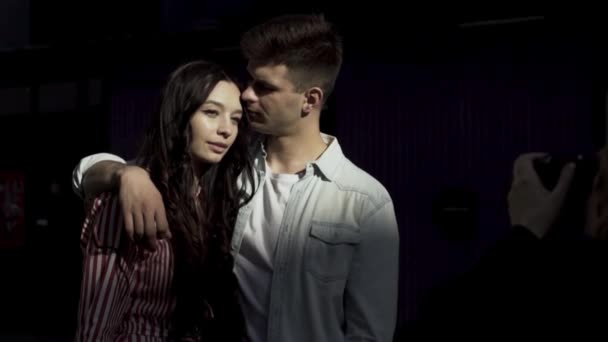 A portrait of a loving caucasian couple while photosession on black background. Action. Caring man hugging a brunette long curly haired woman in a summer dress while standing in front of camera. - Záběry, video