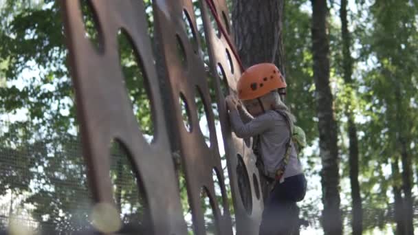 Children in helmets pass the obstacle course in the amusement park in the summer on the trees - Materiał filmowy, wideo