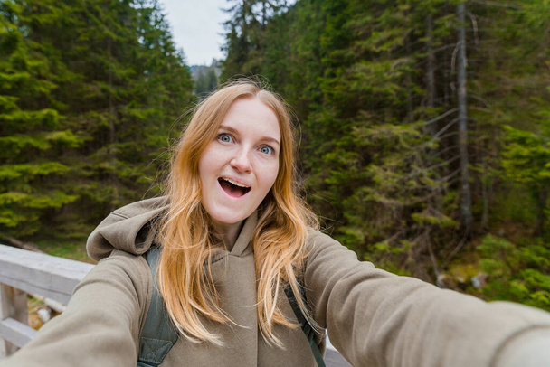Όμορφη νεαρή πεζοπόρος που βγάζει σέλφι στο ορεινό δάσος. Κοιτάζοντας την κάμερα και χαμογελώντας. Ταξίδι και ενεργή ζωή έννοια. Εξωτερικές πόρτες - Φωτογραφία, εικόνα