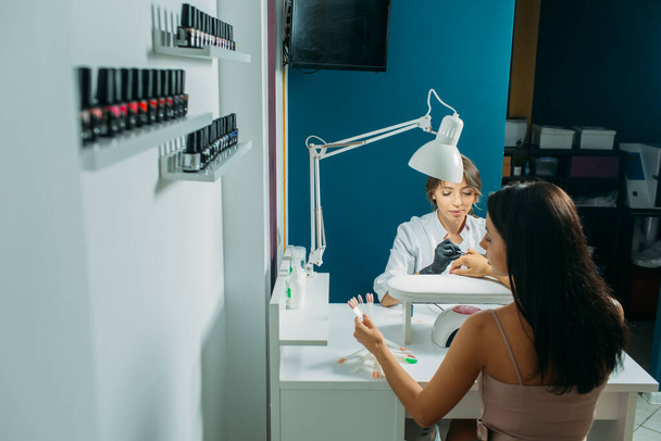 Nail salon, the master does nail care for the client. a master in a dressing gown and a mask, in the frame of the hand, moments of action with the client's nails - 写真・画像