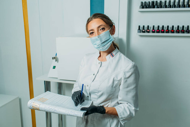 the bright room looks like a medical laboratory, there is an apparatus for quartzing and processing objects in the room. in the frame, a girl in a mask and a medical gown looks very much like a doctor makes an examination and writes in a journal - Fotografie, Obrázek