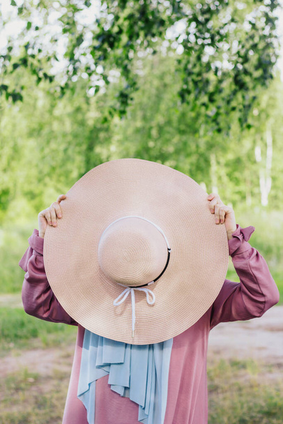 Muslim woman with a hat in a pink dress and a blue hijab with a picnic basket on a background of trees in summer. Modern muslim woman. Muslim woman in summer outfit. Ramadan. - Фото, изображение