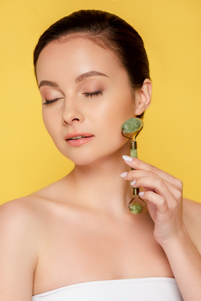 beautiful woman with closed eyes using jade roller on face isolated on yellow - 写真・画像