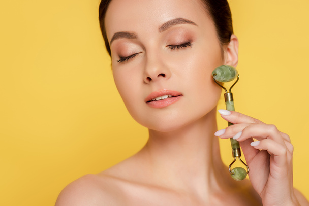 beautiful naked woman with closed eyes using jade roller on face isolated on yellow - Foto, immagini