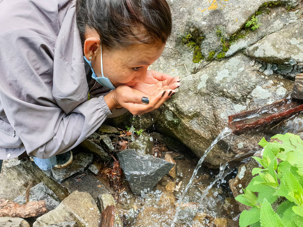 Kilitleme sırasında yüz maskesi takan genç bir Asyalı kızın fotoğrafı. Doğal bir su kaynağında su içiyor. - Fotoğraf, Görsel