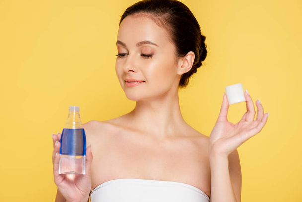 smiling beautiful woman holding micellar water in bottle isolated on yellow - Fotografie, Obrázek