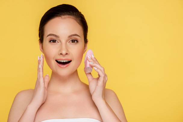 shocked beautiful woman holding facial cleansing brush isolated on yellow - 写真・画像