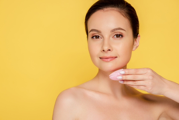 mooie naakte vrouw met behulp van gezichtsreiniging borstel geïsoleerd op geel - Foto, afbeelding