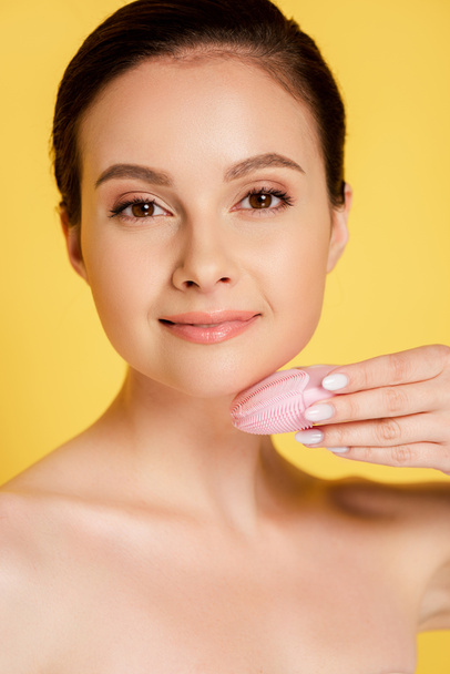 mooie naakte vrouw met behulp van gezichtsreiniging borstel geïsoleerd op geel - Foto, afbeelding