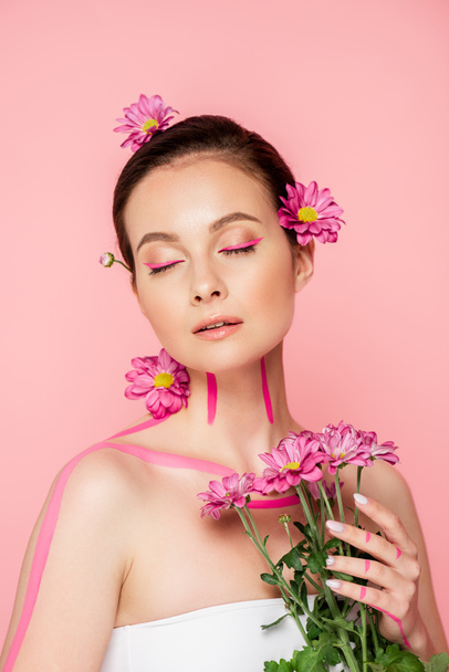 beautiful woman with closed eyes, pink lines on body and flowers in hair holding bouquet isolated on pink - Foto, immagini