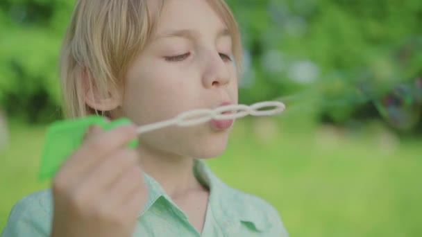 Close-up face of charming blond Caucasian boy blowing soap bubbles outdoors. Portrait of cute little kid having fun in green sunny summer park. Leisure, childhood, joy, lifestyle. - Filmati, video