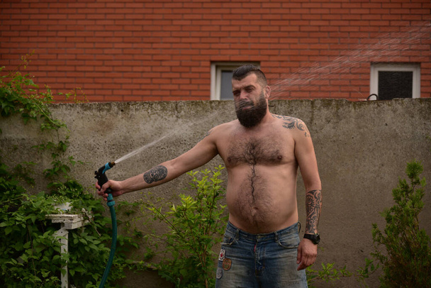 A man douses himself with water from a hose in the street. A man with a beard is watering. A guy in denim shorts. Summer, heat, water - Photo, Image