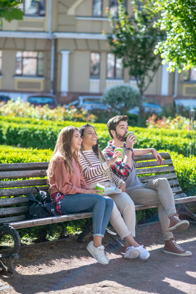Three young friends sitting on bench in yard drinking coffee - Foto, imagen