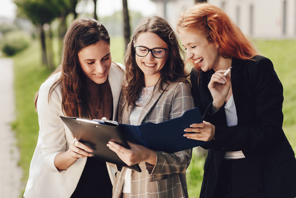 Close up portrait of three successful caucasian businesswomen in suits standing on the street to discuss business. Young women look at documents, talk to each other - Photo, Image
