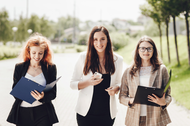 Work outside the office. Three successful caucasian businesswomen in suits standing on the street to discuss business. Young women look at documents, talk to each other - 写真・画像